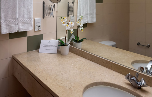 A modern bathroom vanity with a light-colored stone countertop features a sink with a unique faucet, a large mirror reflecting a toilet, and is decorated with a potted orchid and white towels hanging on the wall.