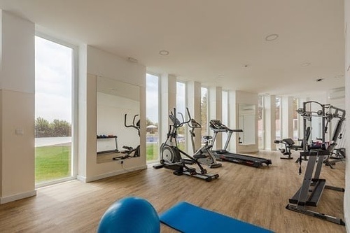 A bright and modern home gym features cardio and weight training equipment, large windows with a view of a lawn, and light wood flooring.