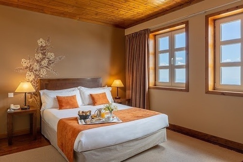 A cozy bedroom features a bed with white and orange linens, a breakfast tray, and two windows offering a glimpse of the outside, all under a wooden ceiling.