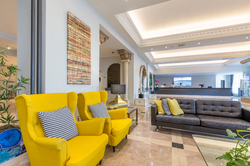O lobby moderno de um hotel apresenta poltronas amarelas vibrantes, um sofá de couro preto e piso de mármore, com a recepção e uma pintura abstrata colorida ao fundo.