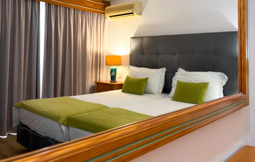 A neatly arranged bedroom featuring a bed with a dark grey tufted headboard, white bedding, and lime green accents is partially reflected in a large wooden-framed mirror in the foreground.
