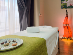 Uma sala de spa ou massagem com uma maca arrumada, um prato decorativo com pedras e uma luminária laranja que cria um ambiente acolhedor e tranquilo.