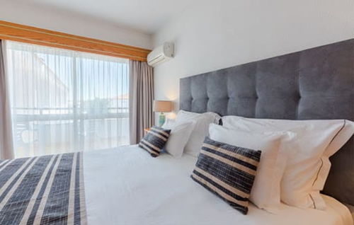 A bright, modern bedroom features a bed with white bedding, striped pillows, and a dark grey tufted headboard, set against a wall with a window revealing sheer curtains and a balcony view.
