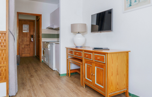 A compact and brightly lit room features a small kitchenette to the left, a wooden console with a wall-mounted TV on the right, and a wooden door in the background.