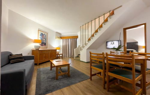 This image shows a cozy, multi-functional living space featuring a sofa and coffee table on a rug, a wooden buffet with lamps, a dining table with chairs, and a staircase leading to an upper level, with a TV mounted beneath it.