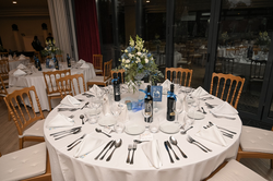 A formal dining room is set with multiple round tables draped in white, each adorned with elegant place settings, blue and white floral centerpieces, and bottles of wine, ready for a special event.