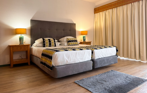 A bright bedroom features two beds with patterned throws and pillows, a gray tufted headboard, and wooden nightstands with lit lamps, alongside light-colored curtains and a small gray rug on a wooden floor.