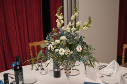 A beautiful white and blue floral centerpiece sits on a formally set dining table with wine bottles, against a backdrop of red curtains.