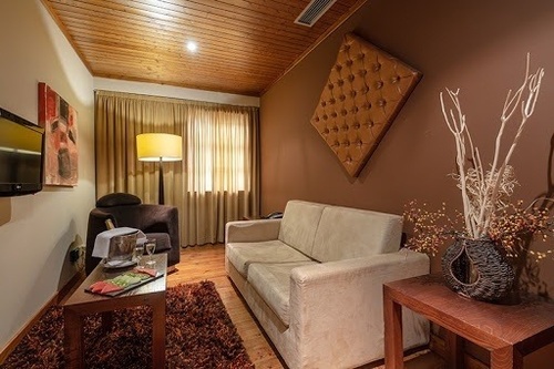 This cozy, earth-toned living space features a light-colored sofa, a dark armchair with champagne on a coffee table, and various decorative accents including a large tufted wall panel and a vase with branches, all beneath a wooden slatted ceiling.