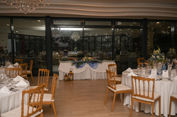 This image captures a beautifully set event room, possibly for a wedding or reception, featuring multiple tables adorned with white linens, chairs, floral arrangements in shades of blue and white, and large windows reflecting chandeliers and the interior, creating an elegant atmosphere.