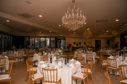This image captures a beautifully set banquet hall adorned with multiple round tables, chairs, and elegant chandeliers, ready for a formal event.