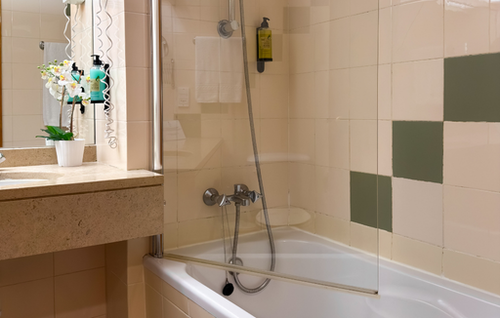 A brightly lit bathroom features a white bathtub with a glass shower screen, tiled walls with light and olive green squares, and a vanity area with a mirror and a potted orchid plant.