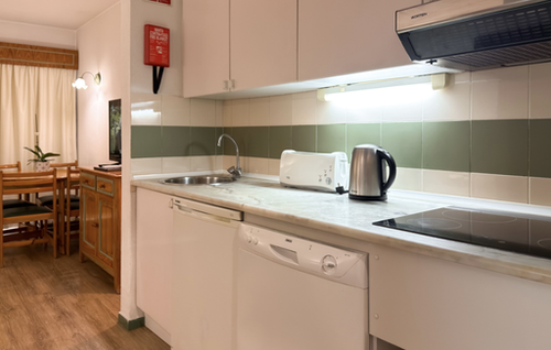 A brightly lit kitchen area with a countertop, sink, and various appliances is shown next to a dining area with a wooden table and chairs.