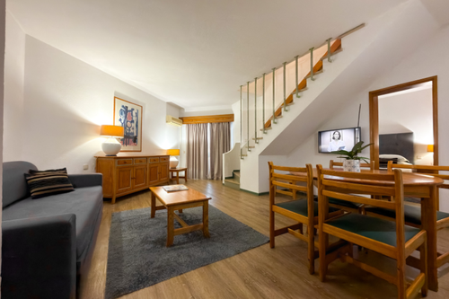 Uma sala de estar e jantar em estilo duplex, mobiliada com sofá cinza, móveis de madeira, tapete e escada interna, com um espelho refletindo um quarto.