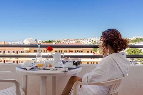 A woman in a white robe enjoys breakfast on a sunny balcony overlooking a town.