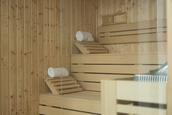 O interior de uma sauna moderna é visível, com paredes e bancos em madeira clara, duas toalhas brancas enroladas sobre apoios de cabeça e um aquecedor de sauna parcialmente visível à direita.