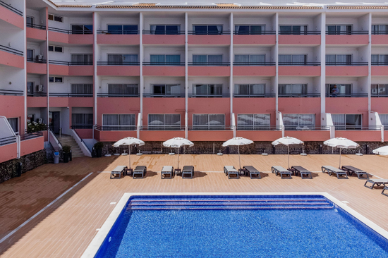 An outdoor swimming pool with sun loungers and white umbrellas sits in the foreground of a multi-story hotel building with pink and white balconies.