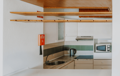This image captures a clean and modern kitchen featuring white cabinets, a tiled backsplash, a sink, an induction hob, a microwave, and overhead wooden shelving, all illuminated by bright ambient light.