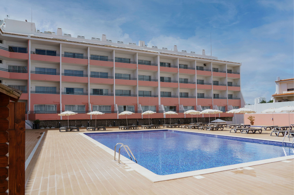 A multi-story hotel or apartment building with pink and white facades and numerous balconies overlooks a bright blue swimming pool surrounded by sun loungers and umbrellas.