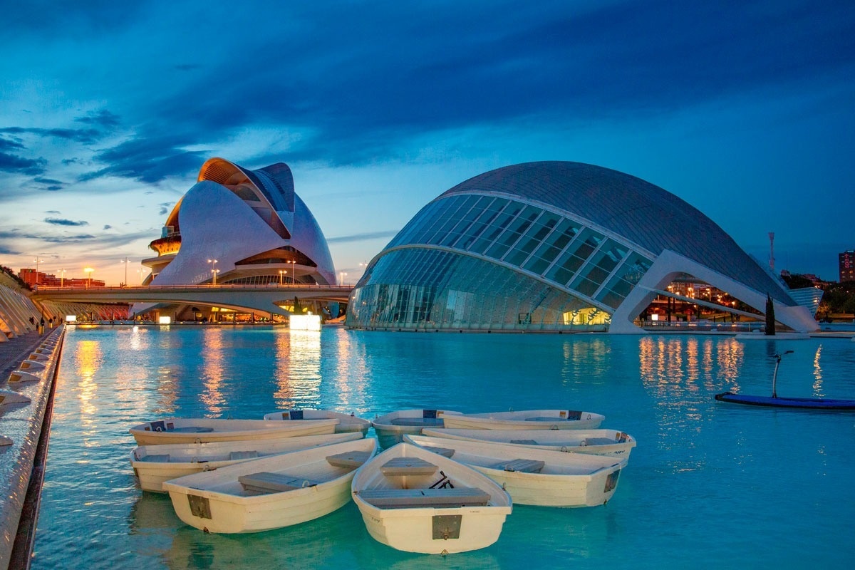 La Ciudad de las Artes y las Ciencias de Valencia se ilumina al anochecer, con sus distintivos edificios reflejándose en las aguas tranquilas donde flotan varias barcas.