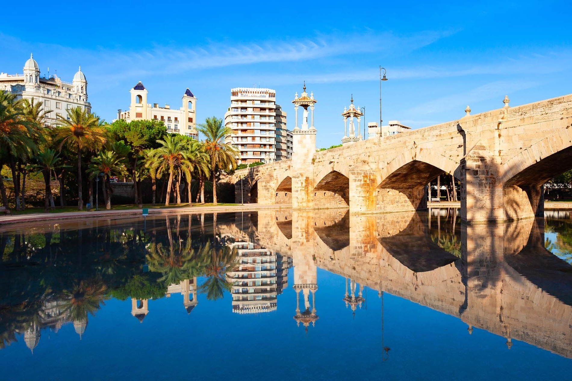 Un puente de piedra con arcos y edificios de la ciudad se reflejan en las tranquilas aguas azules de un canal, rodeados de palmeras bajo un cielo despejado.