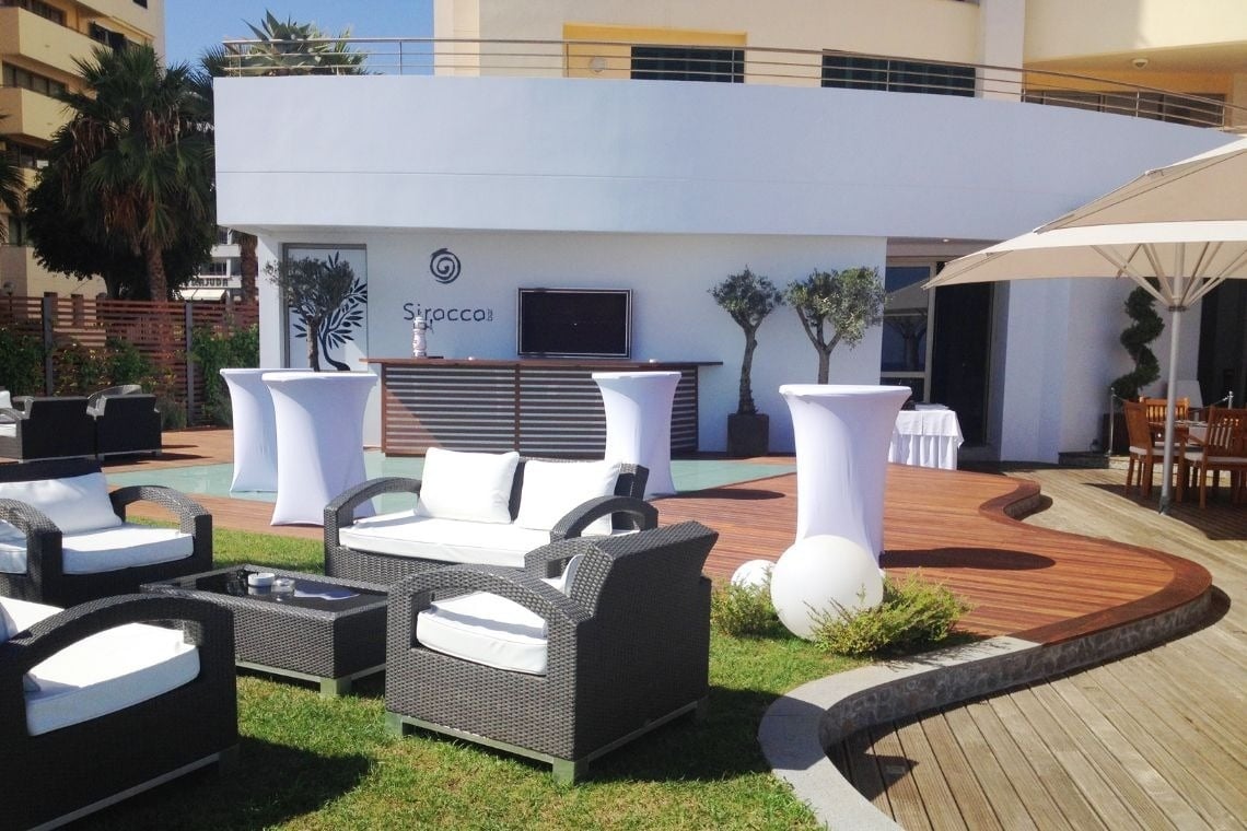 uma área de estar com móveis e mesas em frente a um prédio branco com siroccoi