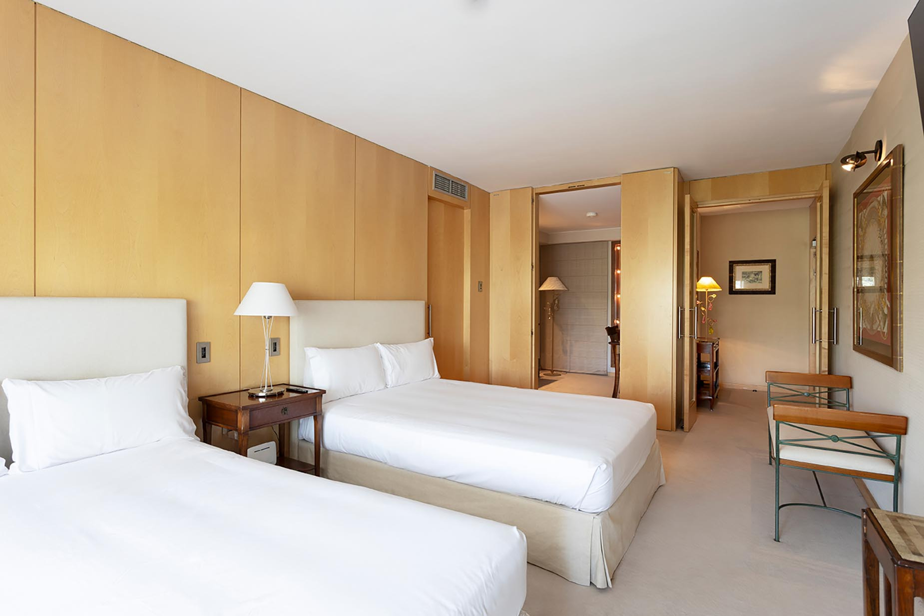 Modern hotel room with two beds, wood paneling, and a glimpse into an adjacent sitting area Suites del Mar Fenals Apartments.