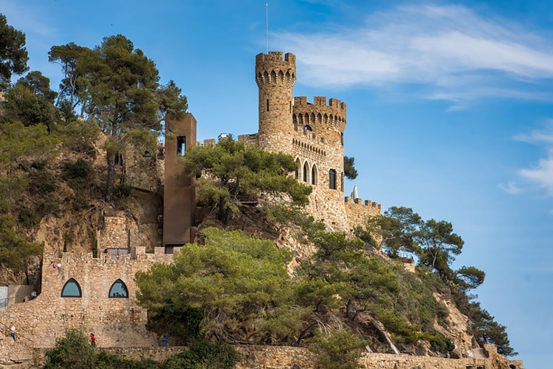 Una vista panoràmica d'una platja costanera amb edificis a la vora, aigua turquesa i roques al primer pla, sota un cel blau amb núvols.