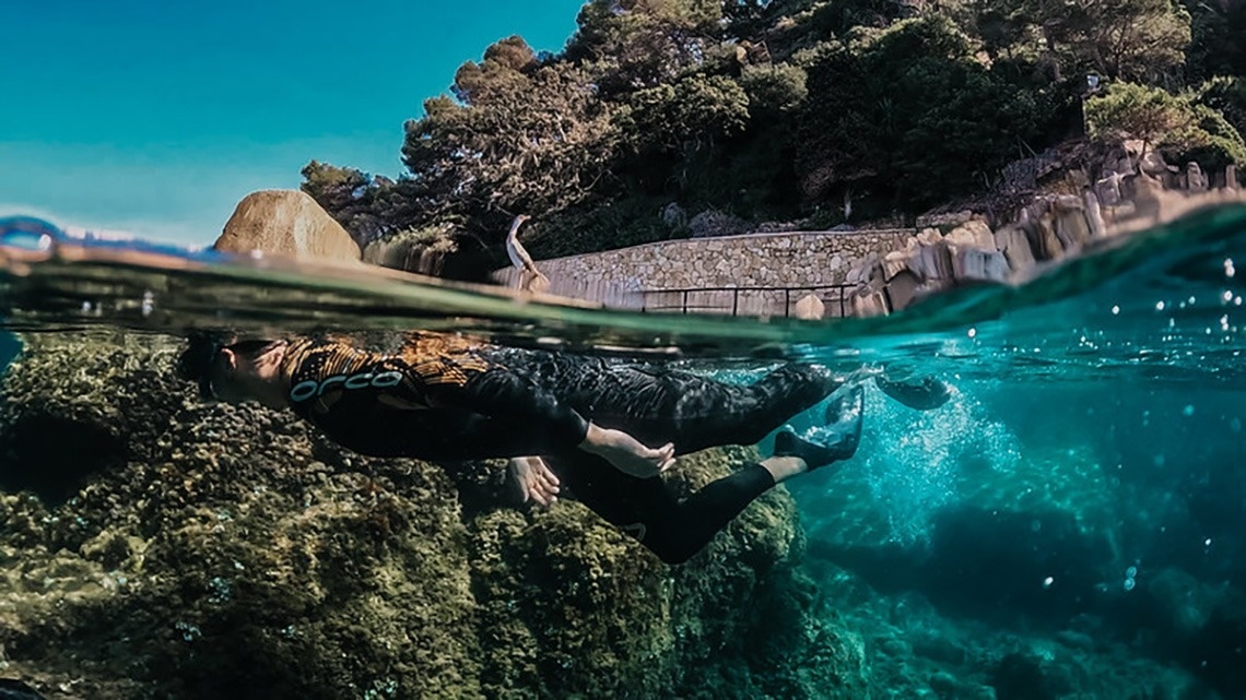 Una vista panoràmica d'una platja costanera amb edificis a la vora, aigua turquesa i roques al primer pla, sota un cel blau amb núvols.
