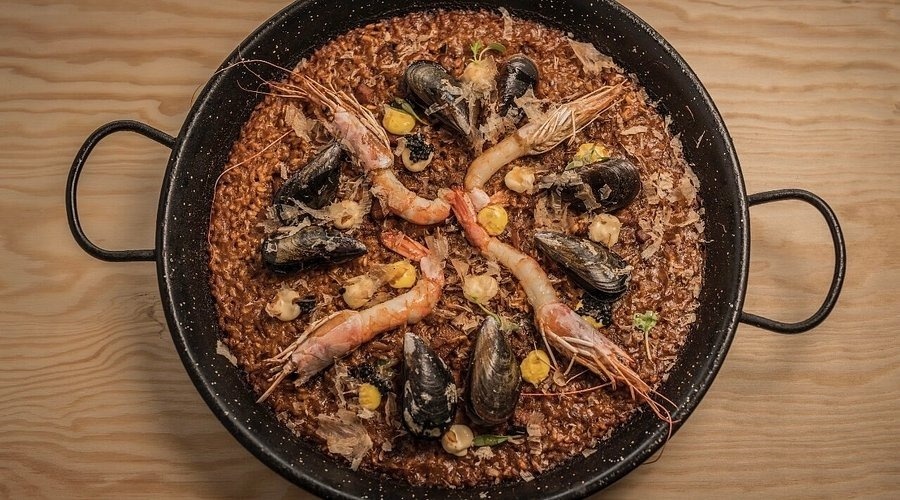 Una paella de marisc amb arròs fosc, gambes, musclos, punts de salsa groga, caviar i encenalls, servida en una paellera negra sobre una taula de fusta.