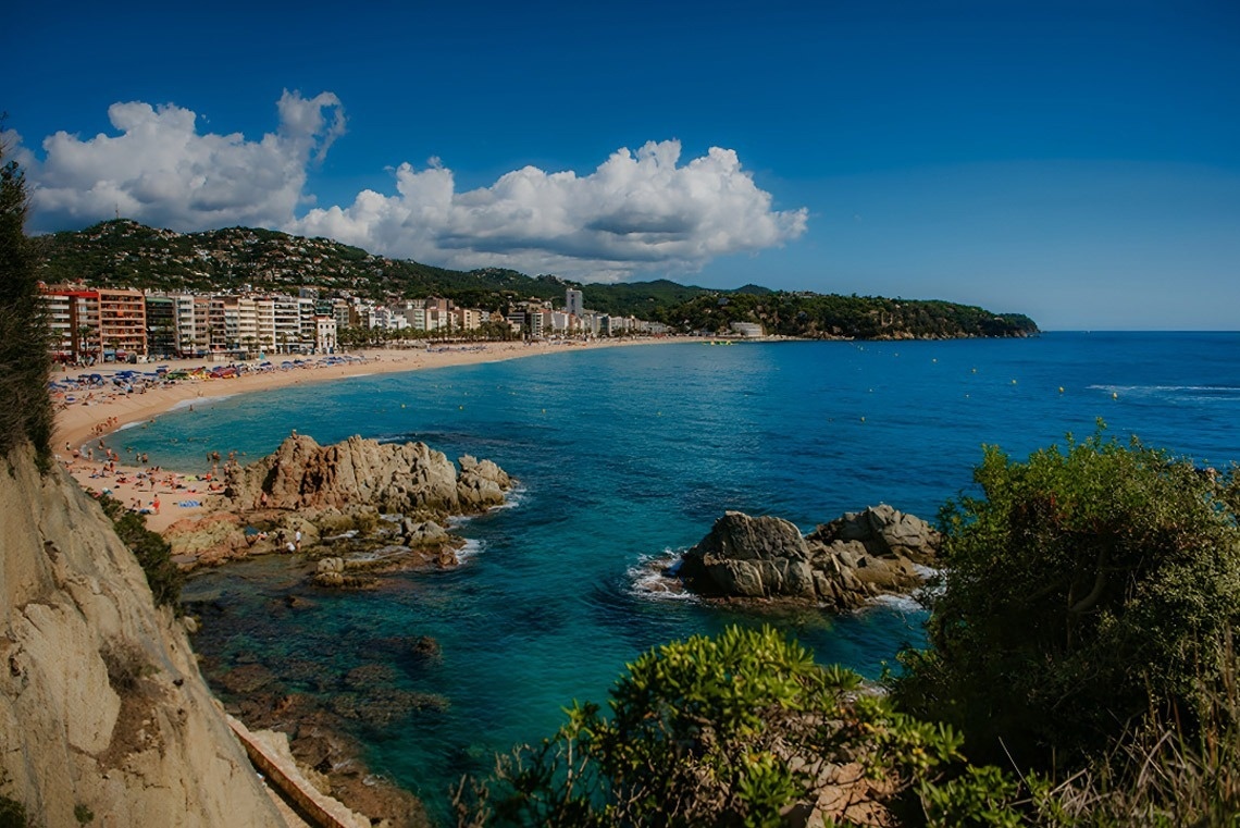 Una vista panoràmica d'una platja costanera amb edificis a la vora, aigua turquesa i roques al primer pla, sota un cel blau amb núvols.