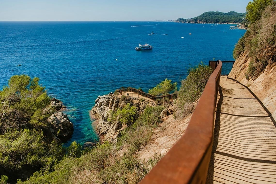 Vista aèria d'una ciutat costanera amb una platja extensa plena de gent, edificis a la vora i turons boscosos, amb nombroses embarcacions a les aigües blaves del mar.