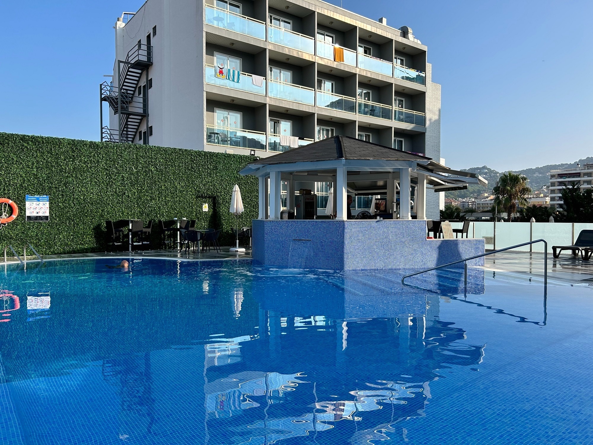 A bright blue tiled swimming pool with reflections of a poolside bar and a multi-story hotel building stands under a clear sky.