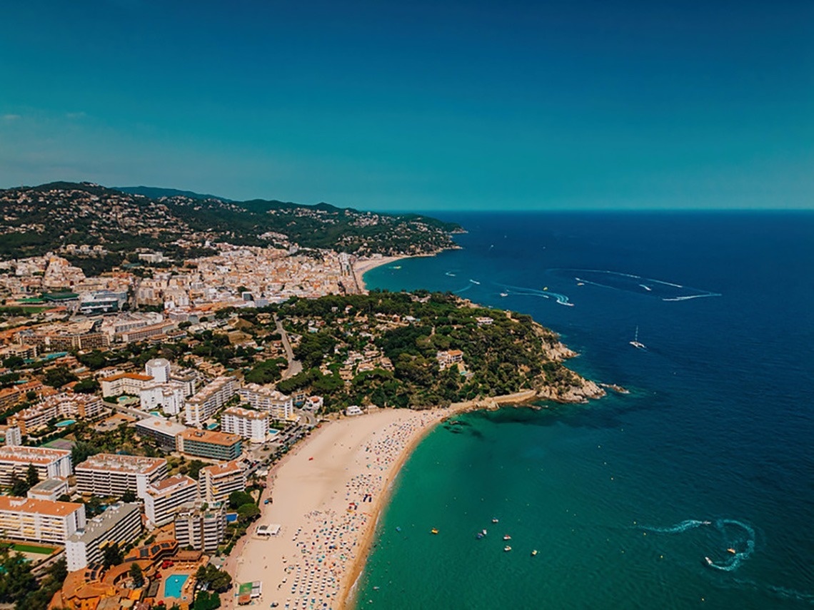 Vista aèria d'una ciutat costanera amb una platja extensa plena de gent, edificis a la vora i turons boscosos, amb nombroses embarcacions a les aigües blaves del mar.