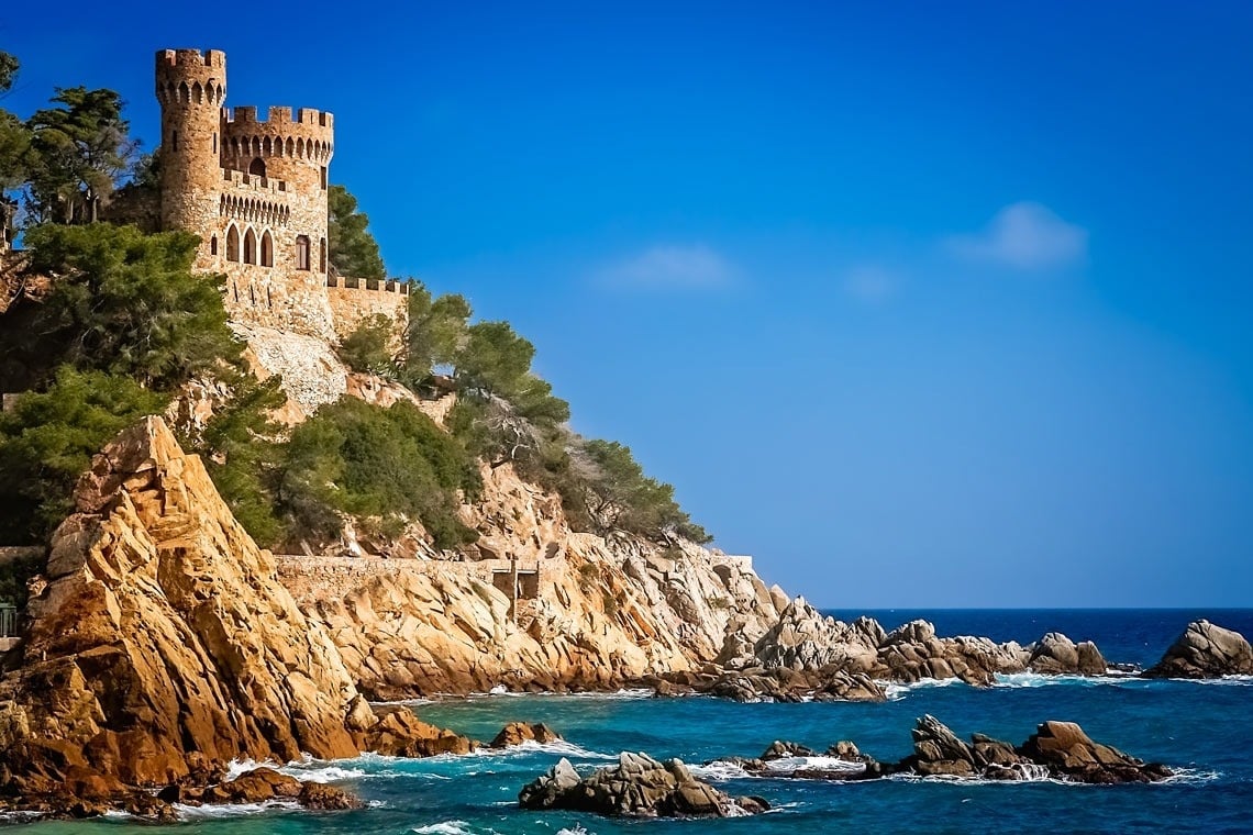 Una costa rocosa i exuberant amb pins s'obre cap a un mar blau intens sota un cel clar, amb algunes barques a la distància.
