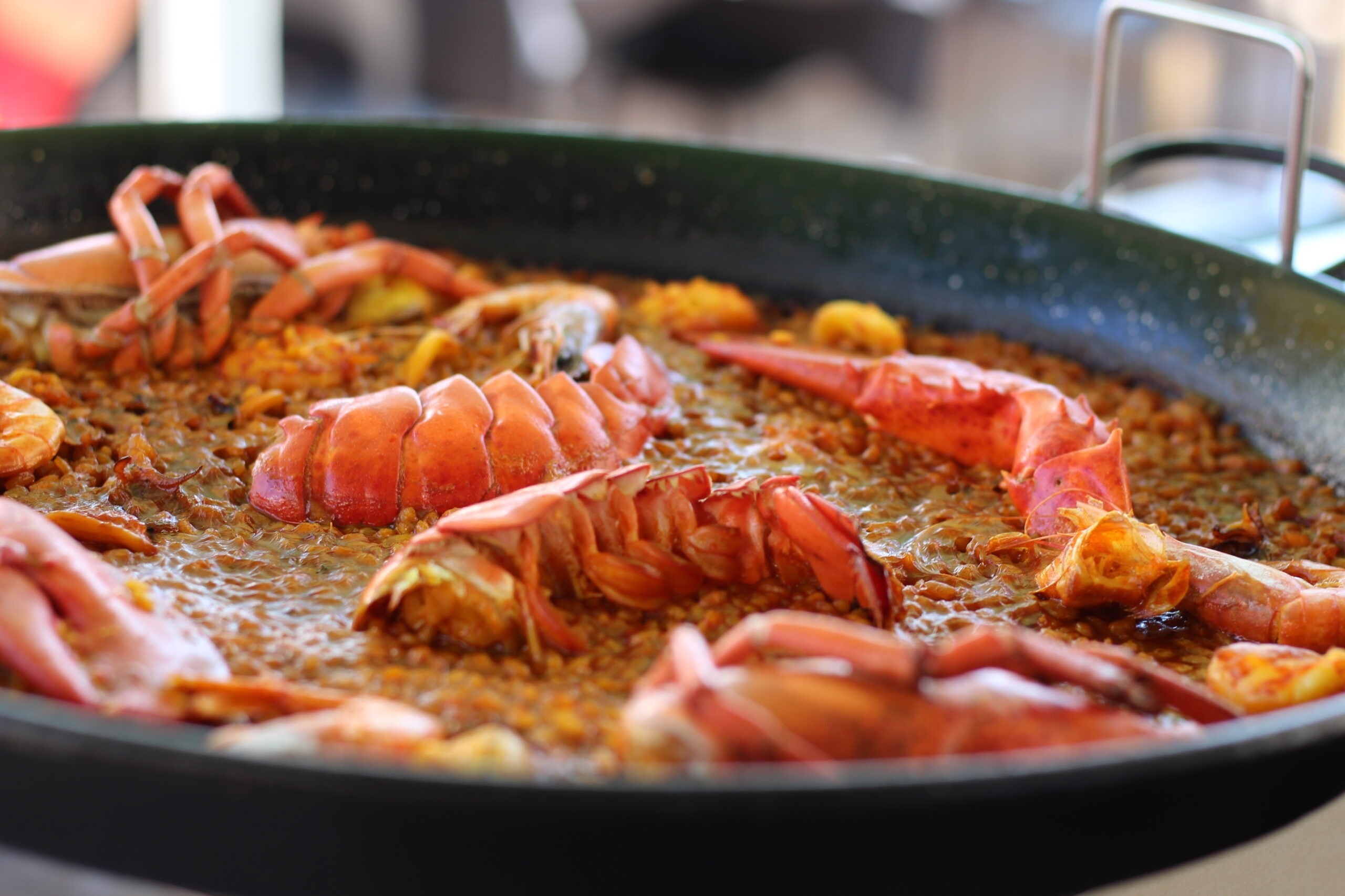 Una paella de marisc amb arròs fosc, gambes, musclos, punts de salsa groga, caviar i encenalls, servida en una paellera negra sobre una taula de fusta.
