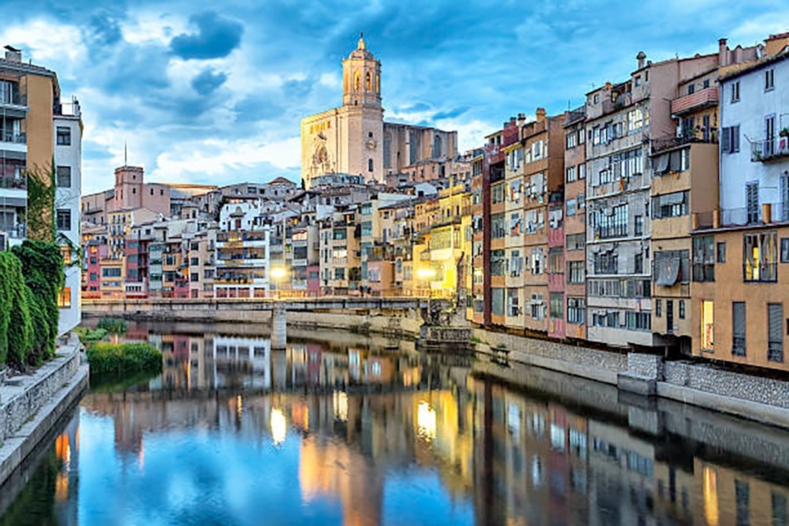 Una vista panoràmica d'una platja costanera amb edificis a la vora, aigua turquesa i roques al primer pla, sota un cel blau amb núvols.