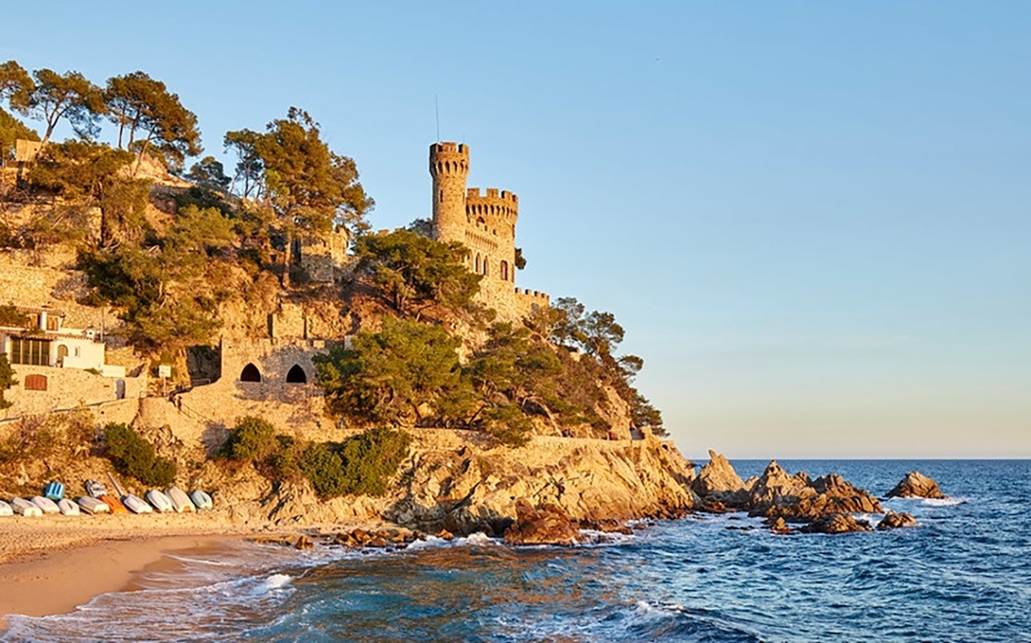 Vista aèria d'una ciutat costanera amb una platja extensa plena de gent, edificis a la vora i turons boscosos, amb nombroses embarcacions a les aigües blaves del mar.