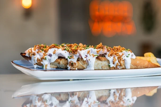 Un llarg entrepà amb salsa blanca, ceba fregida cruixent i cibulet, servit en un plat de vora blava i reflectit a la superfície brillant.