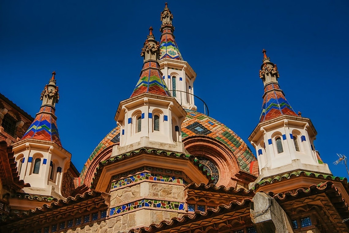 Una vista panoràmica d'una platja costanera amb edificis a la vora, aigua turquesa i roques al primer pla, sota un cel blau amb núvols.
