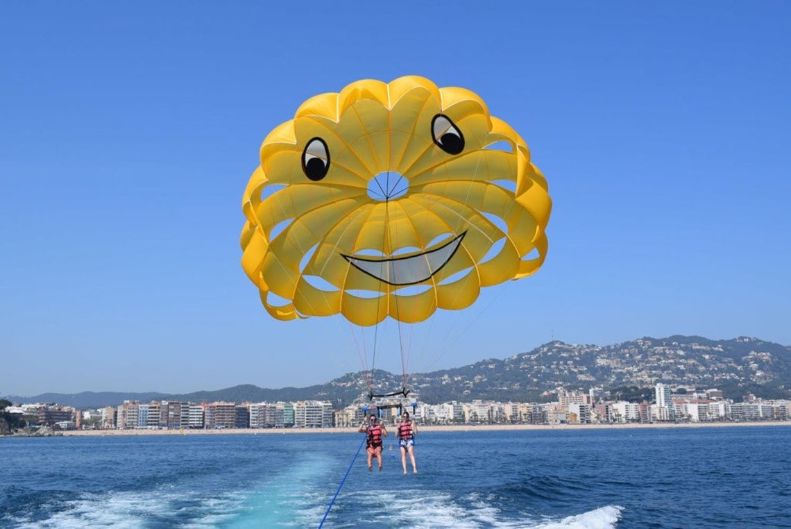 Vista aèria d'una ciutat costanera amb una platja extensa plena de gent, edificis a la vora i turons boscosos, amb nombroses embarcacions a les aigües blaves del mar.