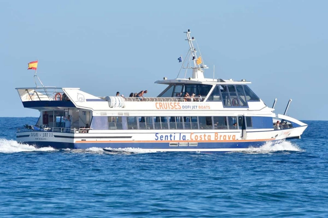 Vista aèria d'una ciutat costanera amb una platja extensa plena de gent, edificis a la vora i turons boscosos, amb nombroses embarcacions a les aigües blaves del mar.