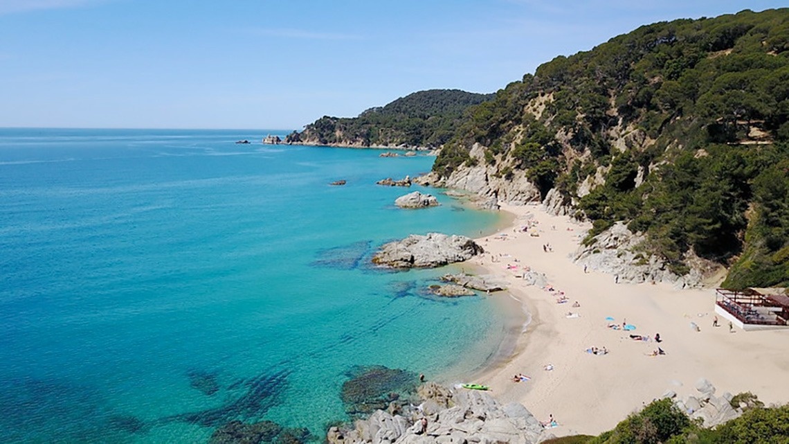 Vista aèria d'una ciutat costanera amb una platja extensa plena de gent, edificis a la vora i turons boscosos, amb nombroses embarcacions a les aigües blaves del mar.