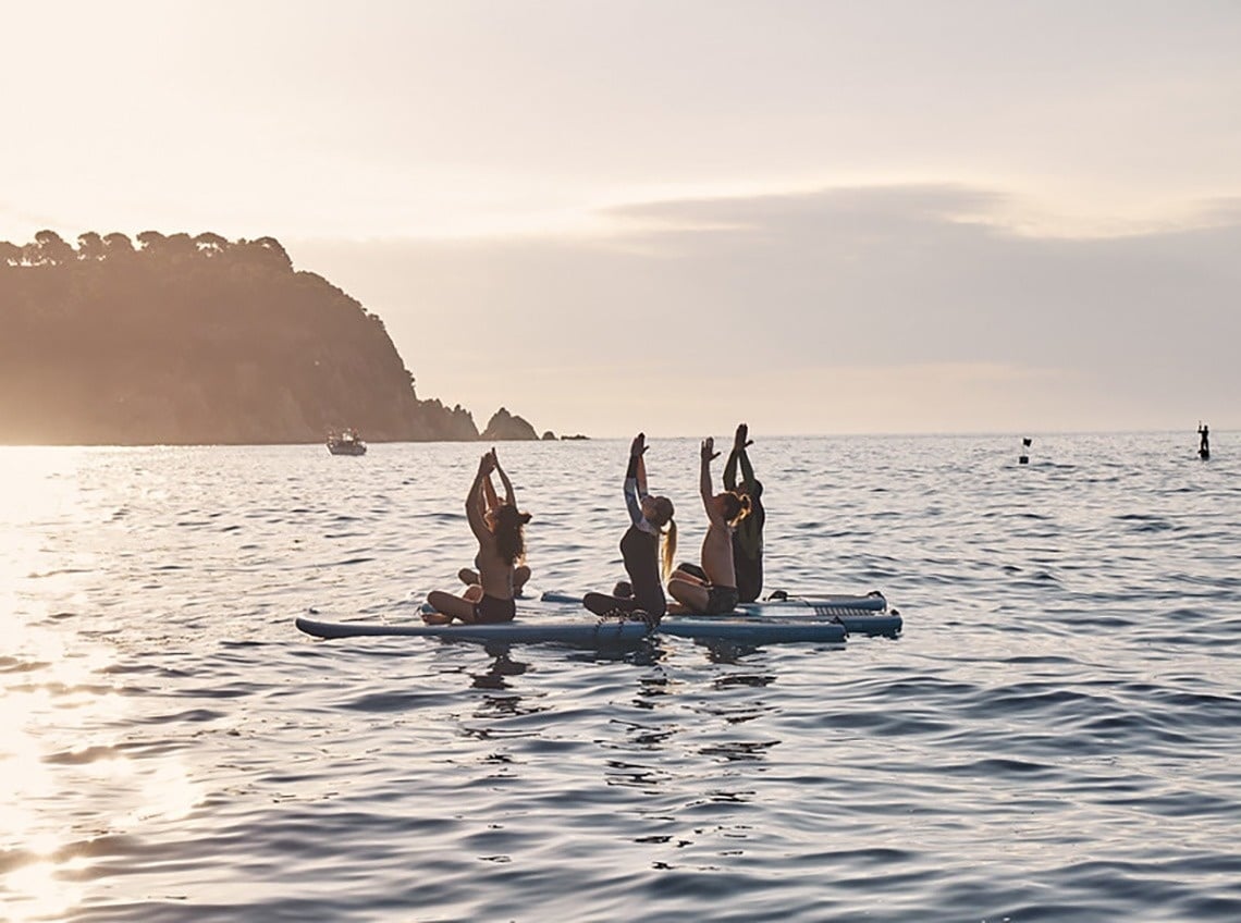 Una vista panoràmica d'una platja costanera amb edificis a la vora, aigua turquesa i roques al primer pla, sota un cel blau amb núvols.