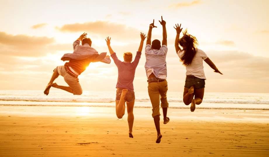 Four energetic people are captured mid-jump on a sandy beach against a vibrant sunset sky, with golden light illuminating their joyful silhouettes.