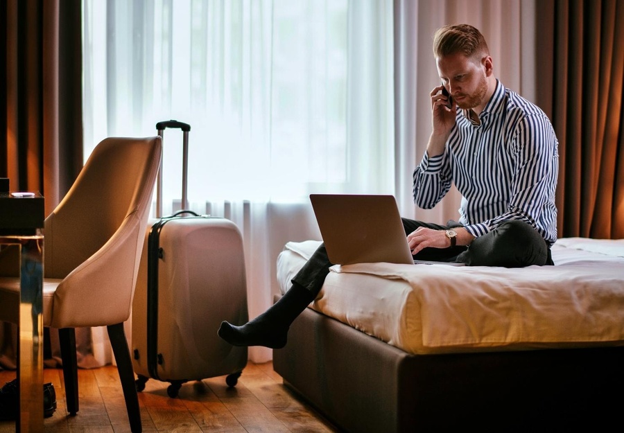a man sits on a bed using a laptop and talking on a cell phone