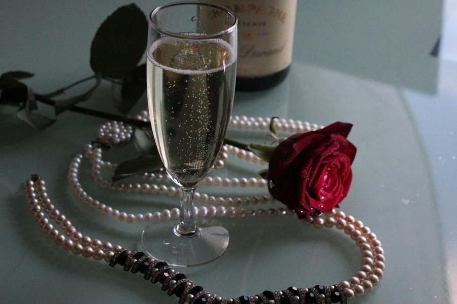 a glass of champagne sits next to a pearl necklace and a rose