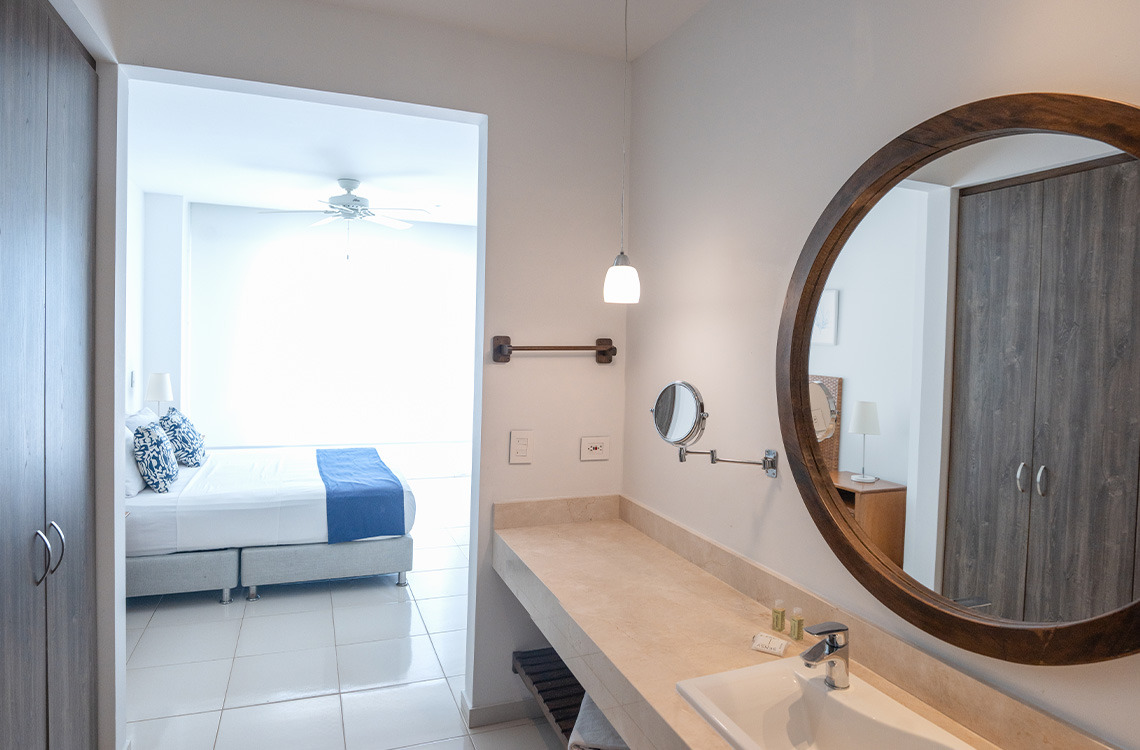 Baño de hotel moderno con espejo circular, tocador y vista a una habitación con cama y ventilador de techo. Marina Puerto Velero