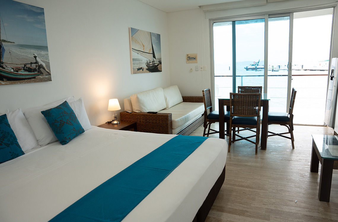 Habitación de hotel con vista al mar y balcón, cama confortable y zona de comedor, perfecta para tu Marina Puerto Velero.