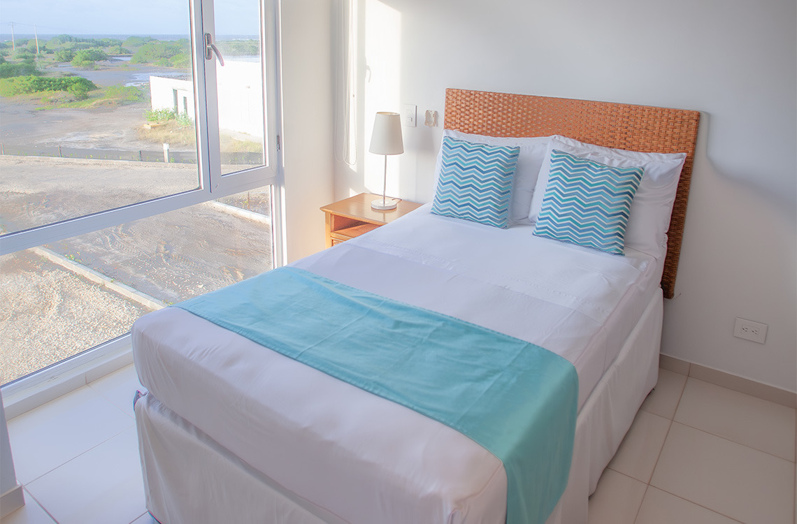 Cama matrimonial con cabecero de mimbre y Marina Puerto Velero, decorada con cojines y manta turquesa, junto a una ventana grande.